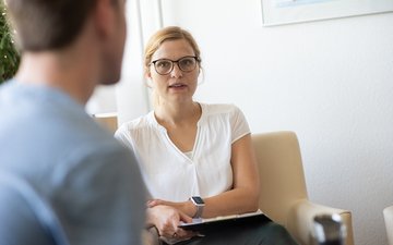 Burnout - Eine Frau im weißen Oberteil mit schwarzer Brille, die auf einer Couch sitzt mit einem Mann im Vordergrund