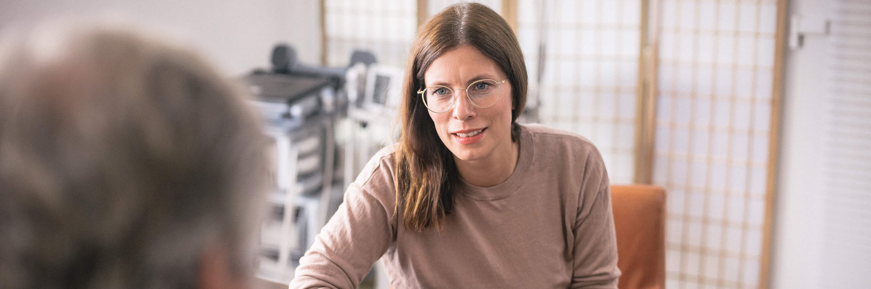 Psychotherapeutin mit braunen Haaren und Brille sitzt an einem Tisch und spricht mit einem Patienten, dessen Rücken zur Kamera zeigt. Im Hintergrund sind medizinische Geräte