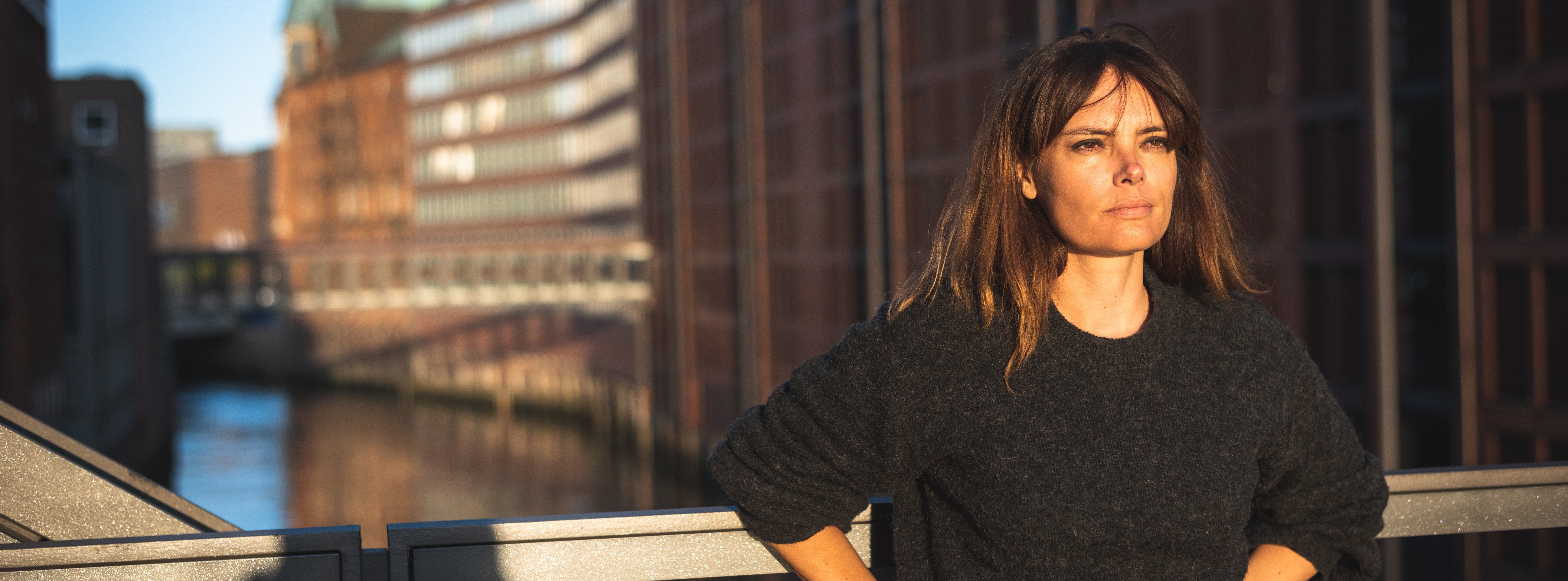 Frau mit schwarzem Pullover und blauer Jeans steht auf einer Brücke und lehnt sich ans Geländer