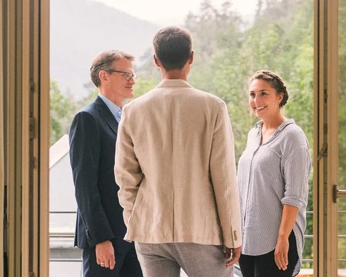 Drei Personen unterhalten sich auf einem Balkon vor einer gr&uuml;nen Berglandschaft