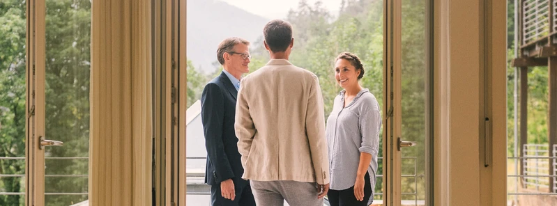 Drei Personen unterhalten sich auf einem Balkon vor einer gr&uuml;nen Berglandschaft