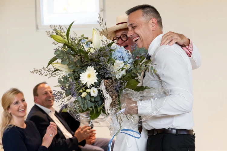 Zwei M&auml;nner umarmen sich bei der Er&ouml;ffnung, einer h&auml;lt einen Blumenstrau&szlig;.