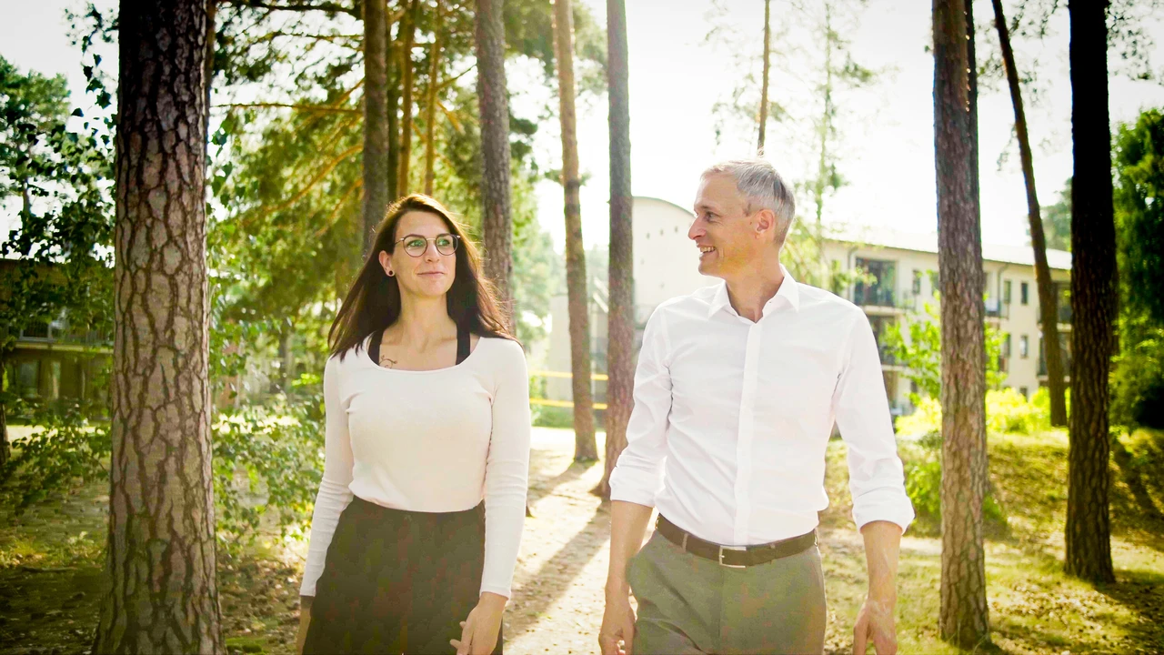 Frau und Mann gehen durch eine gr&uuml;ne Au&szlig;enanlage vor einem Klinikgeb&auml;ude