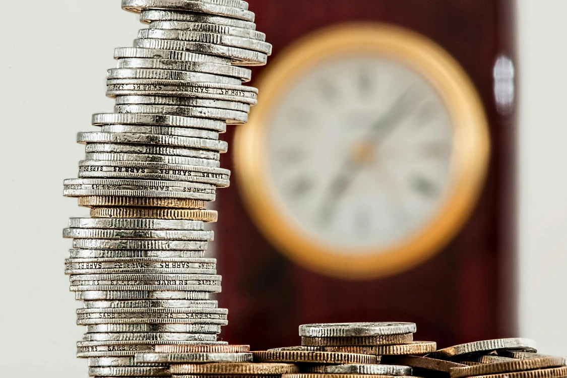 M&uuml;nzstapel vor einer unscharfen Uhr als Symbol f&uuml;r Zeit und Geld