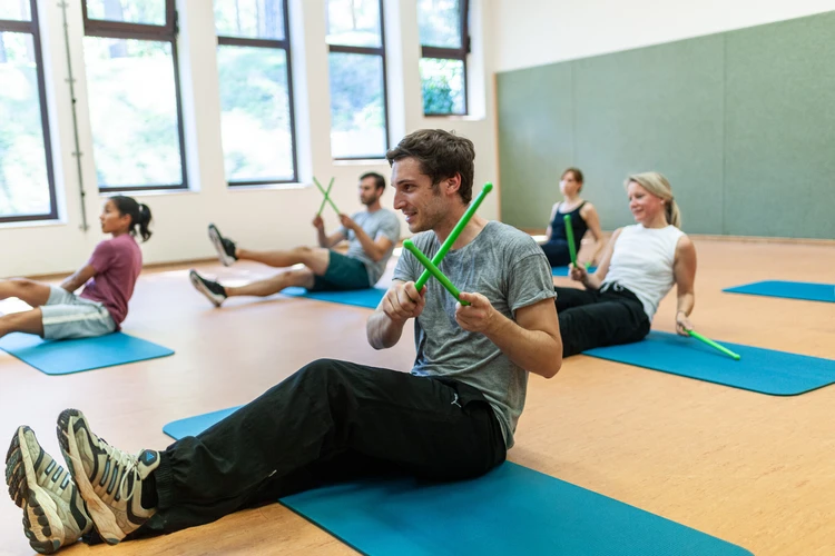 F&uuml;nf Personen sitzen auf Gymnastikmatten und trainieren mit gr&uuml;nen Fitnessst&auml;ben