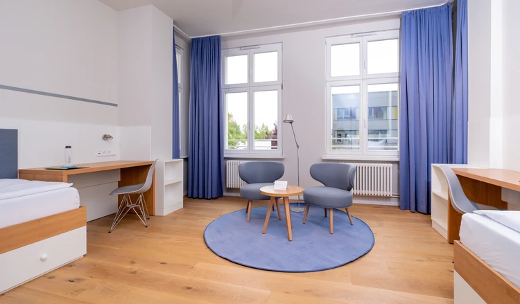 helles Zimmer mit Sesseln, Schreibtischen und Betten