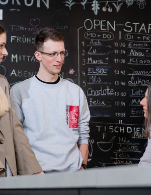 Zwei junge Erwachsene sprechen mit einer sitzenden Frau vor einer Tafel mit Tagesplan