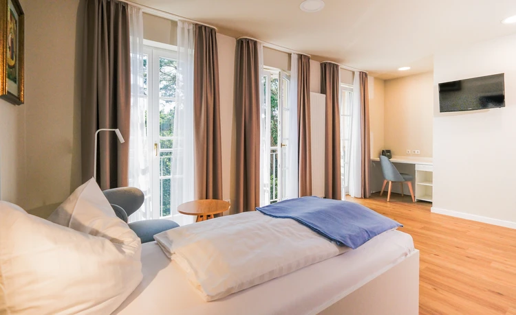 Helles Patientenzimmer mit Bett, Schreibtisch, Fernseher und Balkonfenstern