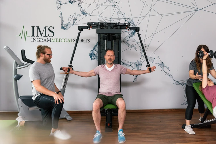 Mann begleitet eine Person bei einer &Uuml;bung im Fitnessraum