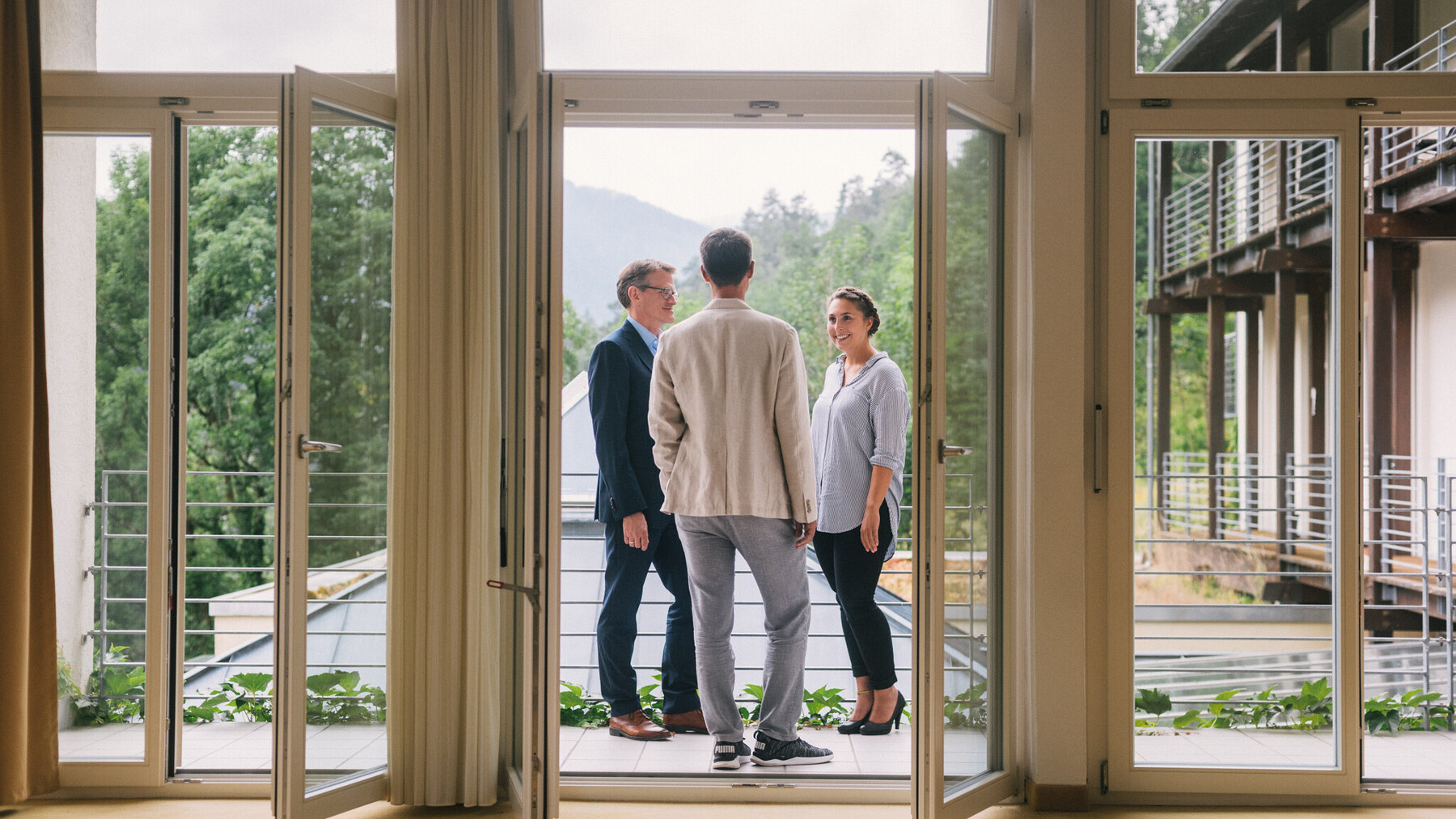 Drei Personen stehen im Gespr&auml;ch auf einem Balkon mit Blick ins Gr&uuml;ne