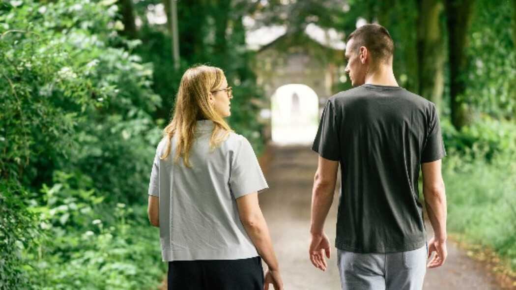 Zwei Personen gehen auf einem von B&auml;umen ges&auml;umten Weg miteinander spazieren