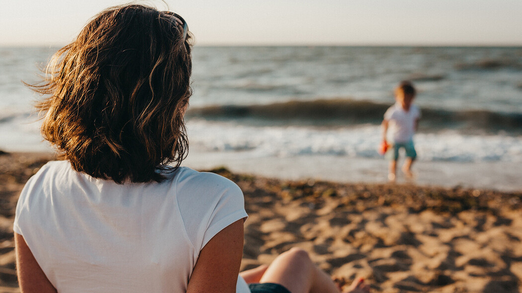 Frau sitzt am Strand und blickt auf ein Kind am Wasser