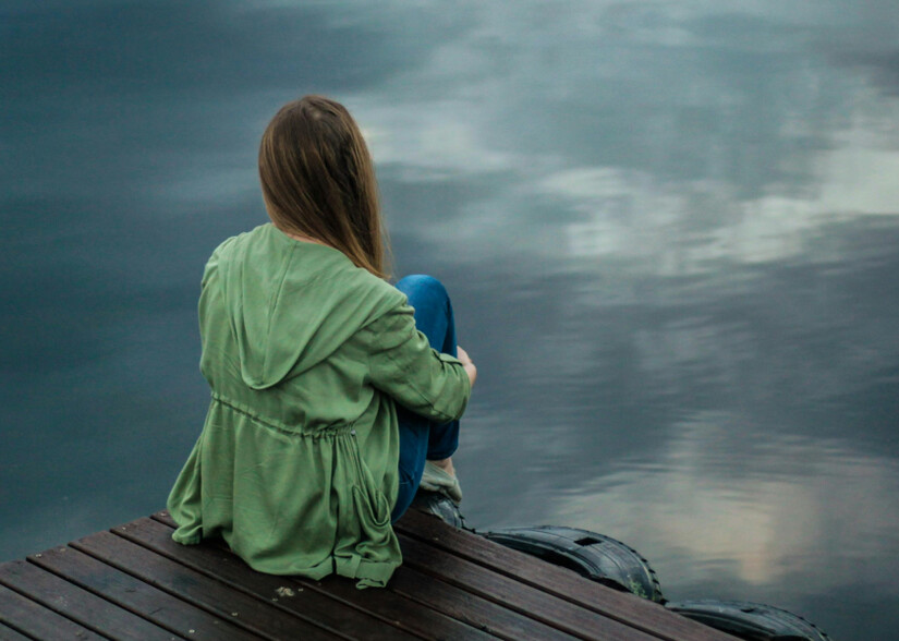 Person sitzt auf einem Steg und blickt auf ruhiges Wasser