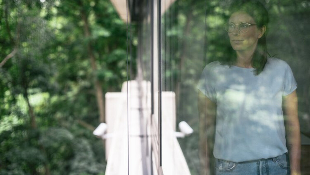 Frau mit Brille steht hinter einer Glasscheibe mit Blick ins Gr&uuml;ne