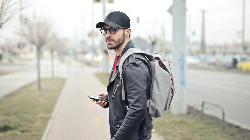 Mann mit Kappe, Rucksack und Smartphone steht auf einem Gehweg