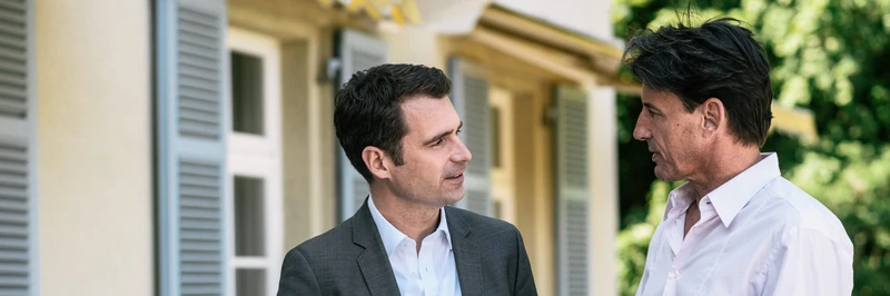 Zwei M&auml;nner stehen vor einem Geb&auml;ude mit Fensterl&auml;den im Gespr&auml;ch